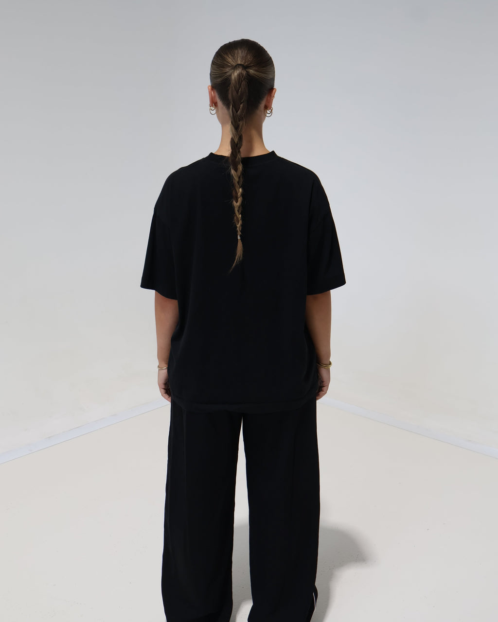 A person viewed from behind wearing a loose black short-sleeve top and matching black wide-leg pants against a plain light gray studio background.