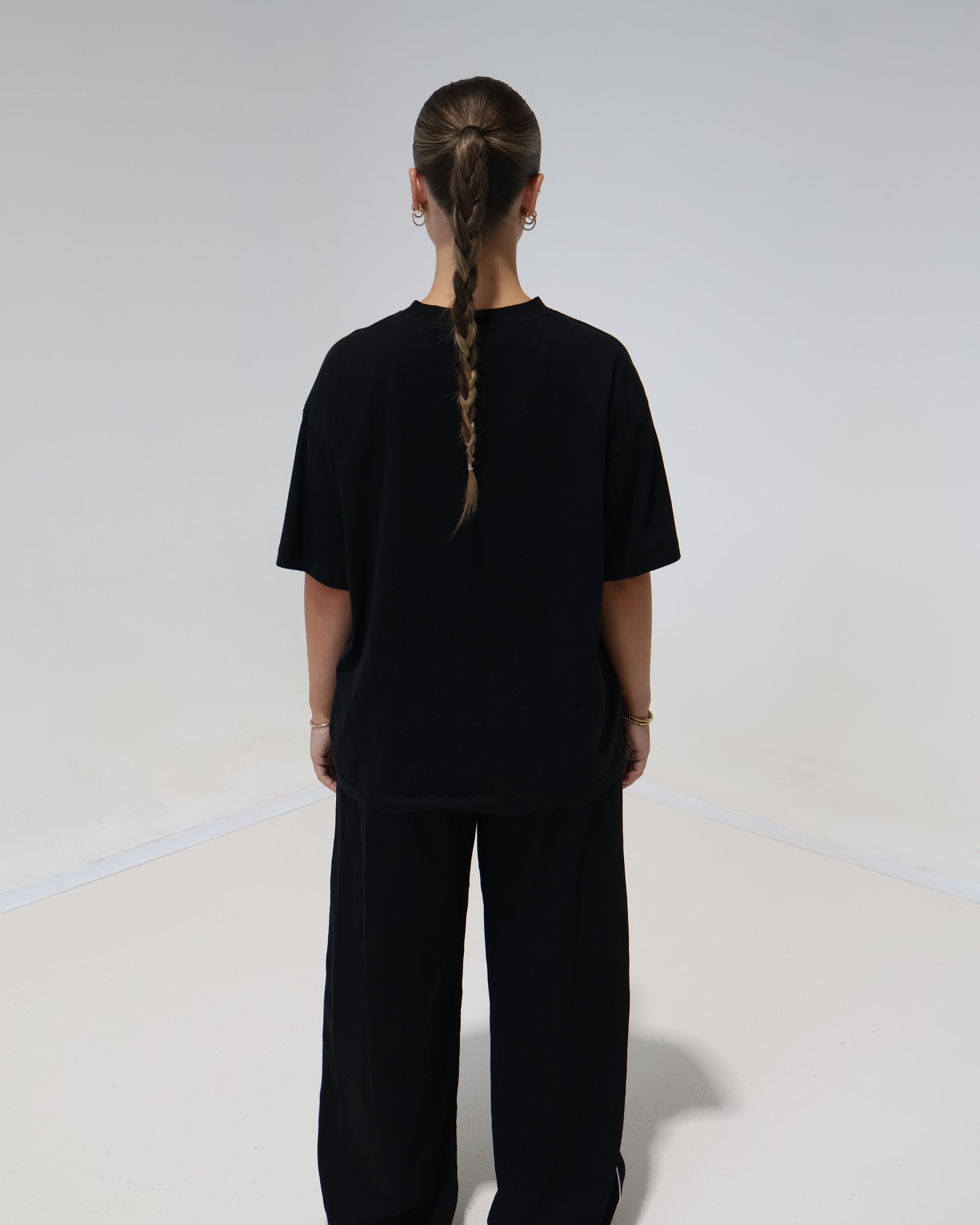 A person viewed from behind wearing a loose black short-sleeve top and matching black wide-leg pants against a plain light gray studio background.